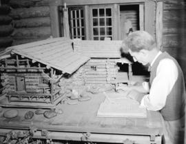 [Man writing in a ledger beside a model log cabin at Silver Creek Camp]