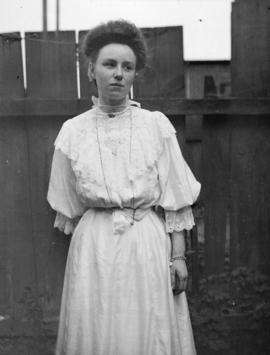 [Young woman in white dress, standing in front of a fence]