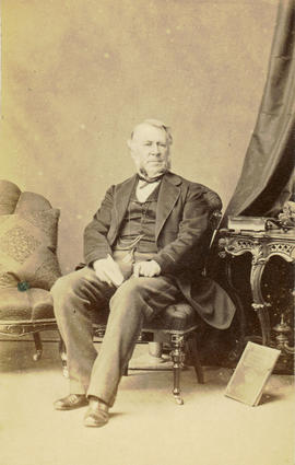 [Seated studio portrait of man, showing chairs and table]