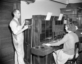 [Man painting a wooden panel beside a switchboard operator at the Mission Telephone Company]