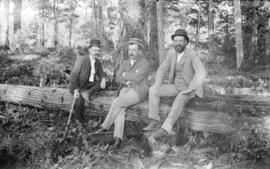 [Three men seated on log in forest]