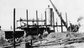 Tank being lifted into place by crane, construction site and workers