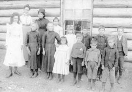 [A teacher and students in front of a log building]