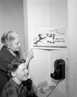 [Woman talking on a public phone while another woman looks at a B.C. Telephone poster]