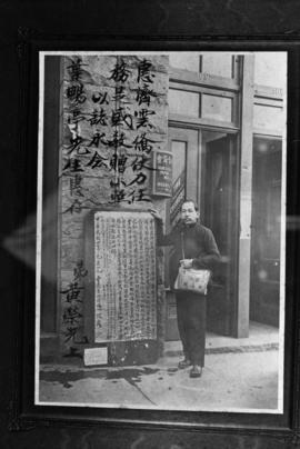Man standing next to a banner and sign outside the Wing Sang Company building at 51 East Pender S...