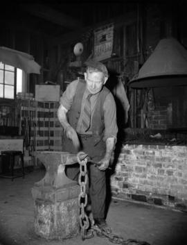 [Blacksmith hammering a chain link on an anvil]