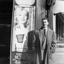 [Joseph Selsey posing beside 'Niagara' poster featuring Marilyn Monroe]