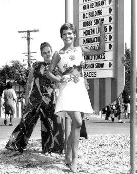 Women in craft dresses pointing at Hobby Show sign on P.N.E. grounds