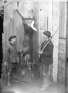 [Man and teenage boy posing with deer strung up in shed]