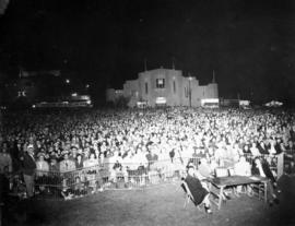 Large audience at Outdoor Theatre stage, with Home Arts building in background