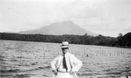 [Man wearing a suit and hat in front of body of water and mountain]