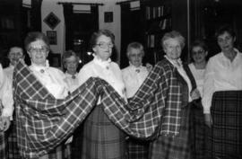 Group of women wearing tartan skirts holding a piece of the Centennial tartan fabric