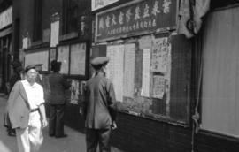 [Men reading Chinese notices and newspapers posted on bulletin boards on Pender Street]