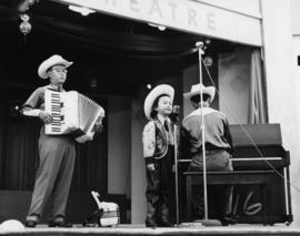 Performance of two youths each playing an accordion and piano with a child dressed in cowboy atti...