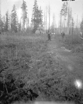 [Two men standing on trail in clearing in front of log cabin]